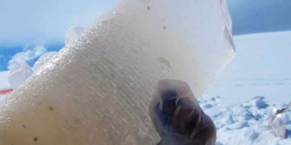 Gloved hand holding a piece of a cylindrical ice core from Hunter, AK outside in the snow. The ice has some small debris and is fairly cloudy at the bottom. Photo by Mike Waszkiewicz.