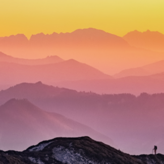 Silhouette of a man hiking along a ridge against a series of overlapping mountain ranges dyed pink by the setting sun.