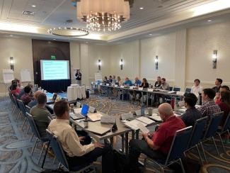 Photograph of Regional Climate Center partners seated around a U-shaped table listening to a presentation being given on a projector screen by a woman. 